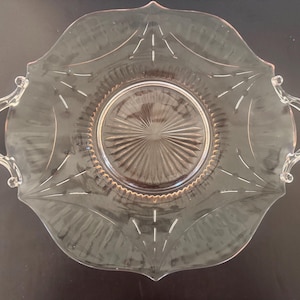 Puede incluir: Un plato de servir de vidrio transparente, de estilo vintage, con borde festoneado y dos asas. El plato presenta un diseño central texturizado y líneas grabadas decorativas que irradian desde el centro. El plato se utiliza probablemente para servir aperitivos o postres.