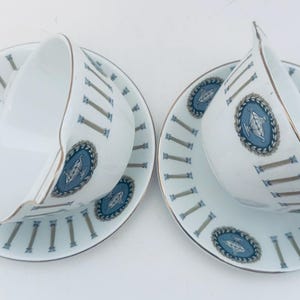 May include: Two white teacups and saucers with a blue and gold design. The saucers have a pattern of vertical lines and a blue medallion. The teacups have a gold trim.