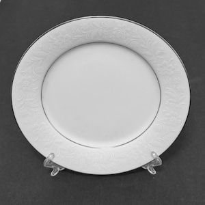 Puede incluir: Un plato de porcelana blanco con un borde floral en relieve y un borde plateado. El plato redondo se encuentra sobre un soporte transparente, sobre un fondo oscuro. El diseño floral en relieve añade un toque de elegancia.