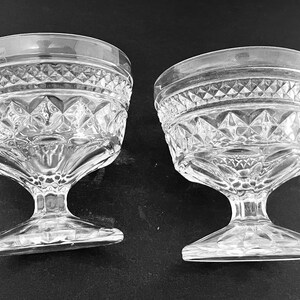 May include: Two clear, stemmed dessert glasses. The glasses have a diamond pattern around the bowl and a decorative rim. The stem is a solid piece of glass that connects to a flat base.