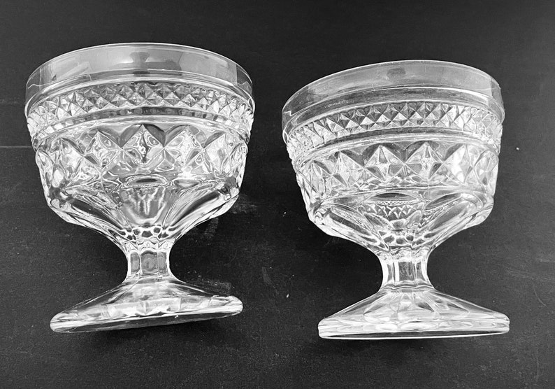 May include: Two clear, stemmed dessert glasses. The glasses have a diamond pattern around the bowl and a decorative rim. The stem is a solid piece of glass that connects to a flat base.