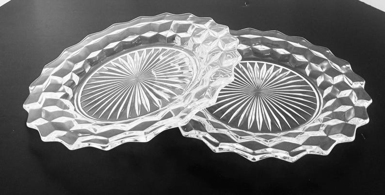 K&ouml;nnte beinhalten: Zwei ovale Schalen aus klarem Glas. Jede Schale hat ein rautenf&ouml;rmiges Muster am Rand und ein Sonnenstrahl-Design in der Mitte. Die Schalen befinden sich auf einer dunklen Oberfl&auml;che.