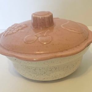 Könnte beinhalten: Ein zweiteiliges Keramik-Serviergeschirr mit Deckel. Der Deckel ist hellrosa mit geprägten Fruchtdesigns, darunter Trauben und Kirschen. Der Boden ist gesprenkelt weiß und hellbraun. Das Geschirr hat einen gewellten Rand und einen Knauf auf dem Deckel.