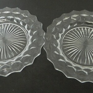 K&ouml;nnte beinhalten: Zwei klare Glasplatten mit einem Rautenmuster am Rand und einem Sonnenstrahl-Design in der Mitte. Die Teller haben einen gewellten Rand und werden auf einer dunklen Oberfl&auml;che pr&auml;sentiert.