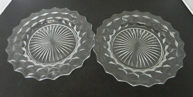 K&ouml;nnte beinhalten: Zwei klare Glasplatten mit einem Rautenmuster am Rand und einem Sonnenstrahl-Design in der Mitte. Die Teller haben einen gewellten Rand und werden auf einer dunklen Oberfl&auml;che pr&auml;sentiert.