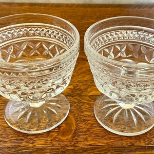 May include: Two clear glass dessert bowls with diamond and starburst patterns. The bowls have a stemmed base and a decorative rim. The bowls are sitting on a wooden surface.
