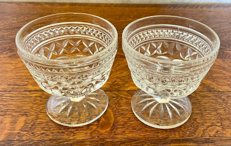 May include: Two clear glass dessert bowls with diamond and starburst patterns. The bowls have a stemmed base and a decorative rim. The bowls are sitting on a wooden surface.