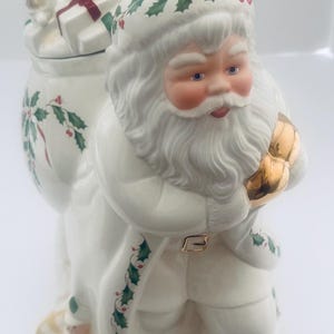 Puede incluir: Figura de cerámica de Papá Noel con abrigo y gorro blancos, adornada con acebo y lazos rojos. La figura lleva un saco lleno de regalos y un cuerno dorado. Papá Noel tiene barba blanca y mejillas rosadas, ideal para la decoración navideña.