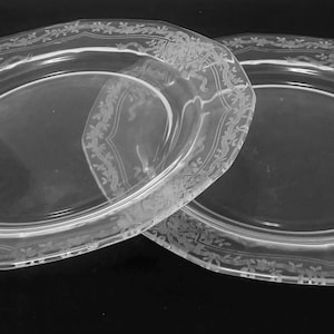 May include: Two clear glass plates featuring an etched floral and ribbon pattern along the scalloped edges. The plates have a smooth, transparent center and a decorative border design that encircles the entire rim of each circular piece.