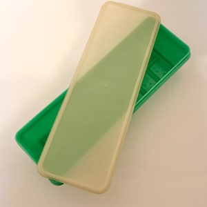 May include: A green rectangular container with a translucent lid. The lid is slightly ajar, revealing the interior of the container. The container is made of plastic and appears to be empty. The lid has a slight tint of yellow.