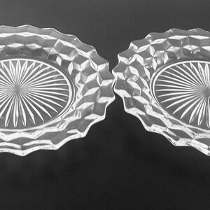 K&ouml;nnte beinhalten: Zwei klare Glasplatten mit rautenf&ouml;rmigem Rand und einem Sonnenstrahl-Design in der Mitte. Die Platten sind oval und liegen auf einer dunklen Oberfl&auml;che. Geeignet zum Servieren von Vorspeisen oder Desserts.