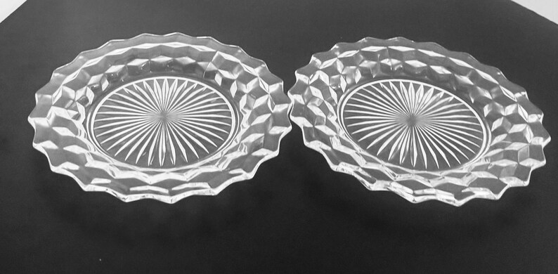 K&ouml;nnte beinhalten: Zwei klare Glasplatten mit rautenf&ouml;rmigem Rand und einem Sonnenstrahl-Design in der Mitte. Die Platten sind oval und liegen auf einer dunklen Oberfl&auml;che. Geeignet zum Servieren von Vorspeisen oder Desserts.