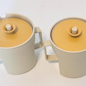 Può includere: Due brocche vintage color crema con coperchi giallo senape. Ogni brocca ha un manico quadrato e un coperchio con un piccolo pomello bianco. Una brocca ha un beccuccio. Le brocche sono probabilmente di plastica.