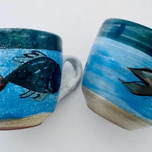 May include: Two ceramic mugs with a hand-painted fish design. The mugs feature a blue and white colour scheme, with a dark blue fish and a brown tail. The mugs have a handle and a slightly textured surface.