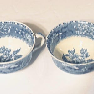 Op de afbeelding: Twee bijpassende blauw-witte porseleinen theekopjes. De kopjes hebben een bloemenontwerp in het midden en een gedetailleerde rand. Elk kopje heeft een oor en een brede, ondiepe kom. De kopjes staan tegen een witte achtergrond.