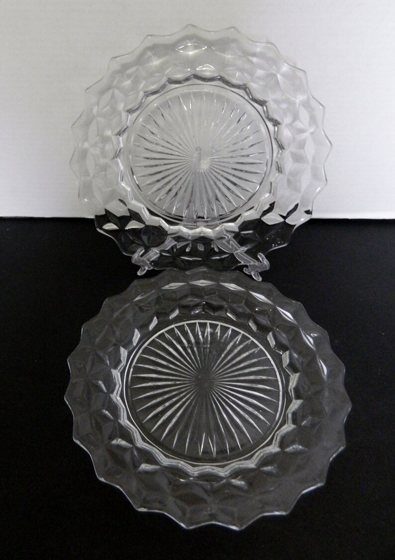 K&ouml;nnte beinhalten: Zwei klare Glasplatten mit einem Diamantmuster an den R&auml;ndern und einem Sonnenstrahl-Design in der Mitte. Die Teller werden auf einer schwarzen Oberfl&auml;che mit wei&szlig;em Hintergrund pr&auml;sentiert.