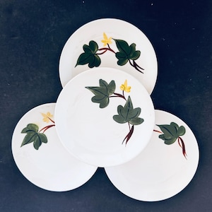 Può includere: Quattro piatti in ceramica bianca con un motivo di foglie verdi e fiori gialli. Le foglie sono verde scuro con steli marroni e fiori gialli. I piatti sono disposti su una superficie blu scuro.