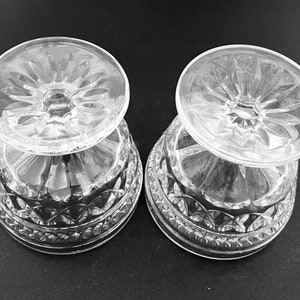 May include: Two clear, cut-glass dessert dishes. The bowls have a diamond pattern around the base and a starburst design on the bottom. The dishes are turned upside down, revealing the intricate details of the glasswork. The background is a solid black.