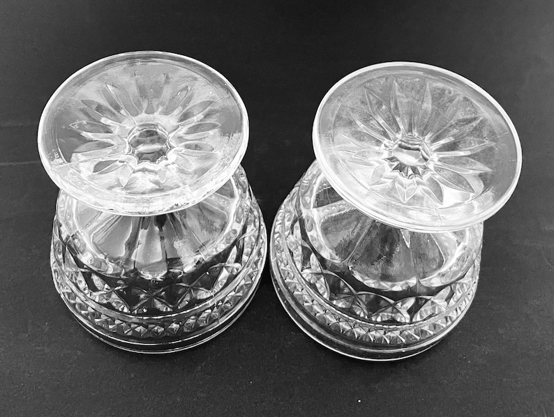May include: Two clear, cut-glass dessert dishes. The bowls have a diamond pattern around the base and a starburst design on the bottom. The dishes are turned upside down, revealing the intricate details of the glasswork. The background is a solid black.
