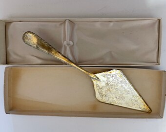 May include: Silver-colored cake server with an ornate floral design on the blade, presented in a beige box with a satin lining. The server has a decorative handle and is likely vintage.