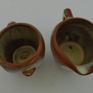 以下が含まれることがあります： クリーマーと砂糖入れの2つのセラミック製サービングピースが表示されています。クリーマーにはハンドルと注ぎ口が付いています。どちらのピースも茶色の縁と白と茶色の内側を持っています。砂糖入れには小さなハンドルが付いています。