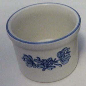 Puede incluir: Un pequeño cuenco de cerámica blanquecina con un diseño floral azul y un borde azul. El cuenco tiene una parte superior ligeramente inclinada y una forma cilíndrica. El diseño floral está en el lateral del cuenco.