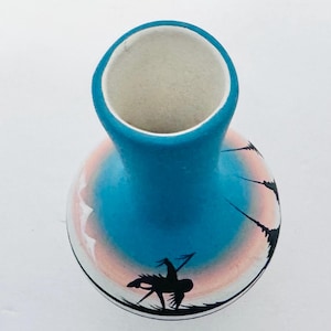 Könnte beinhalten: Eine handbemalte Keramikvase mit blauem Hals und rundem Körper. Der Körper zeigt ein Sonnenuntergangsdesign mit rosa und weißen Farbtönen sowie schwarze Silhouetten von Figuren und Pflanzen. Die Vase hat einen weißen Innenraum.