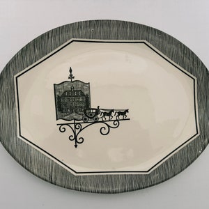 May include: Oval serving platter with a cream-colored center and a black and white textured border. The center features a black illustration of a building, carriage, and horses. The platter has a black geometric border.