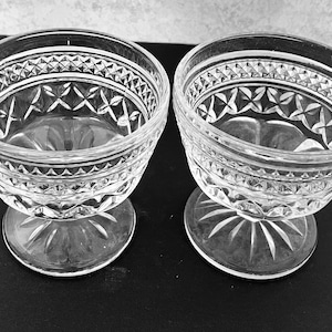 May include: Two clear glass dessert bowls with ornate diamond and star patterns. The bowls have a rounded shape with a pedestal base. The glass has a cut crystal appearance, reflecting light. The bowls are likely vintage or antique.