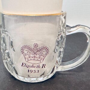 May include: Clear glass beer mug with a handle, featuring a purple crown design and the text "Elizabeth R 1953". The rim of the mug has a gold accent. The mug is partially filled with a white substance.