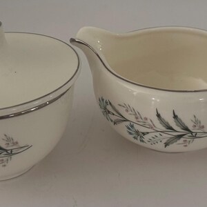 May include: A cream-colored ceramic sugar bowl with a matching lid and a creamer pitcher. Both pieces feature a delicate botanical pattern of blue and grey floral sprigs and leaves, accented with a thin silver rim along the edges of the vessels.