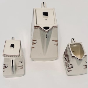 May include: A three-piece white ceramic tea set with brown and black accents. The set includes a teapot with a lid, a sugar bowl with a lid, and a creamer. The set has a geometric design and is suitable for serving tea.