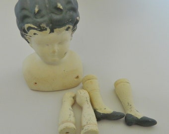 Op de afbeelding: Een vintage poppenhoofd met grijs haar en een crèmekleurig lichaam. De afbeelding toont ook losse poppenarmen en -benen, waaronder twee armen en twee benen met grijze schoenen. De poppenonderdelen zijn verouderd en vertonen tekenen van slijtage.