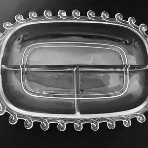 May include: Clear glass serving dish with three compartments and handles. The oval-shaped dish has a decorative scalloped edge. The dish is designed for serving appetizers or snacks.