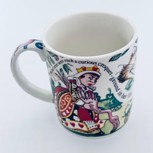 Könnte beinhalten: Weiße Keramik-Tasse mit einer farbenfrohen Illustration der Herzkönigin aus Alice im Wunderland. Die Tasse hat einen gebogenen Henkel und den Text "never seen such a curious croquet-ground in her life."