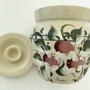 May include: A beige ceramic jar with a lid, hand-painted with red cherries, white blossoms, and green leaves. The jar has a rounded shape and a matching lid. The word "Peace" is written on the jar.