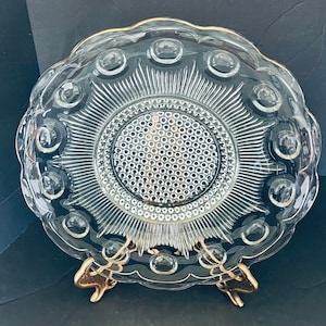 Puede incluir: Plato de servir de vidrio transparente con borde festoneado y ribete dorado. El plato presenta un diseño central texturizado y está sostenido por un soporte dorado. El plato tiene forma circular con un patrón de círculos en relieve.