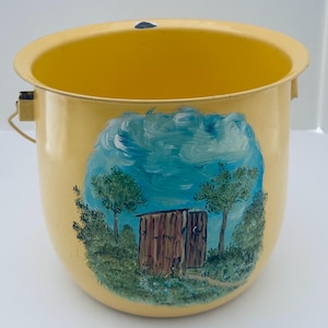 May include: A yellow metal bucket with a hand-painted scene of a wooden outhouse, trees, and a blue sky. The bucket has a handle and a small, dark-colored attachment near the rim.