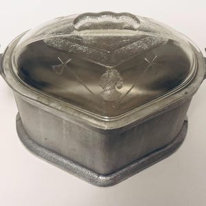 May include: A heart-shaped metal casserole dish with a clear glass lid. The lid features an embossed profile of a person and crossed arrows. The base is made of a matte gray metal with two side handles, and the glass lid has a central handle.