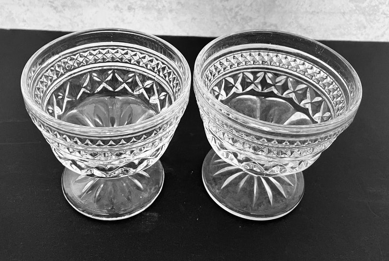 May include: Two clear glass dessert bowls with intricate diamond and starburst patterns. The bowls have a rounded shape with a pedestal base. The glass has a textured, cut-glass appearance, adding to their vintage charm.
