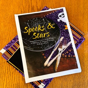 May include: A magazine titled "Spooks & Stars" with a black cover featuring a spiderweb design and the text in yellow. Silverware is on the magazine. The magazine rests on a purple and orange patterned placemat.