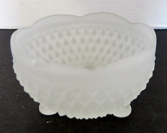 May include: A frosted glass bowl with a scalloped rim and a diamond pattern on the exterior. The bowl has three small feet and a smooth interior. The bowl is white and appears to be made of glass.