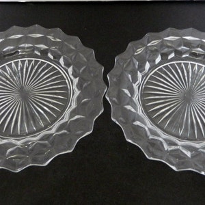 K&ouml;nnte beinhalten: Zwei Dessertteller aus klarem Glas. Jeder Teller hat einen rautenf&ouml;rmigen Rand und eine Mitte mit einem Sonnenstrahl-Design. Die Teller sind oval und liegen auf einer dunklen Oberfl&auml;che.