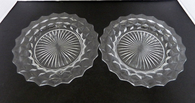 K&ouml;nnte beinhalten: Zwei Dessertteller aus klarem Glas. Jeder Teller hat einen rautenf&ouml;rmigen Rand und eine Mitte mit einem Sonnenstrahl-Design. Die Teller sind oval und liegen auf einer dunklen Oberfl&auml;che.