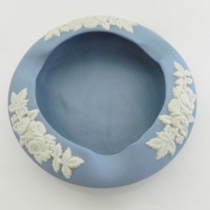 Puede incluir: Un plato redondo de cerámica azul claro con una depresión central. El borde está decorado con patrones de relieve de flores y hojas blancas. El plato tiene un acabado liso y mate sobre un fondo blanco.