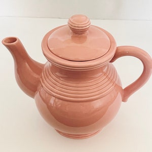 Puede incluir: Una tetera de cerámica rosa polvoriento con tapa y asa. La tetera tiene un cuerpo redondeado con detalles horizontales acanalados y una boquilla curva. La tapa está rematada con un pequeño pomo redondo. La tetera está diseñada para preparar té.