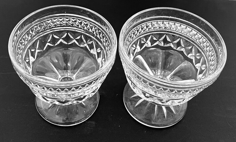 May include: Two clear, vintage-style glass dessert bowls. Each bowl features a decorative diamond and star pattern around the rim and base. The bowls are elevated on a short, stemmed foot.