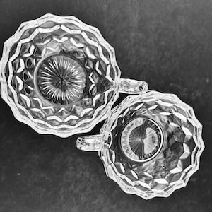 May include: Two clear, faceted glass teacups with handles are arranged on a dark surface. The cups have a scalloped rim and a starburst design in the center. The handles are small and curved.