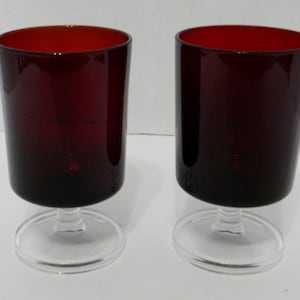 Könnte beinhalten: Zwei Weingläser mit klarem Stiel und Fuß und einer dunkelroten Schale. Die Schalen sind zylindrisch mit leicht ausgestelltem Rand. Die Gläser sind aus Glas und stehen vor einem weißen Hintergrund.