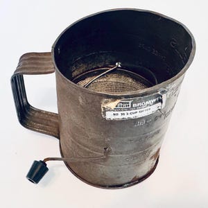 Pode incluir: Peneira de metal vintage com uma alça e uma manivela. A peneira tem uma tela de malha no interior e um rótulo que diz "Leigh Bromwell No. 39 3-Cup Sifter". A peneira é de cor metálica escura e envelhecida.